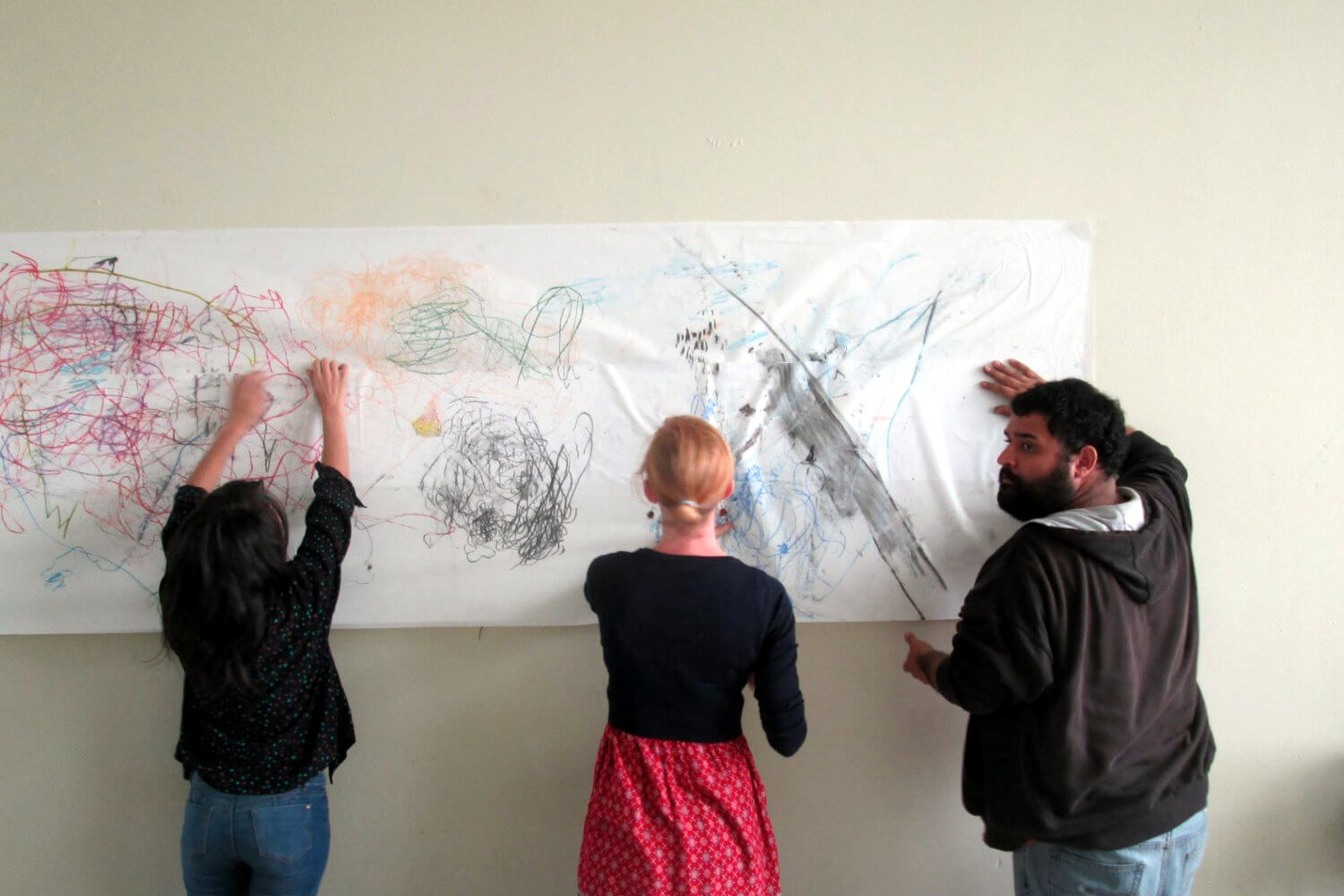 Tres personas, dos mujeres y un hombre, dibujando y pintando sobre un papel blanco montado sobre una pared