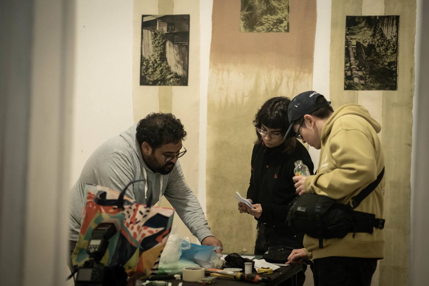 Tres personas, dos hombres y una mujer, trabajando sobre una mesa con múltiples objetos, con telas con imágenes en el fondo