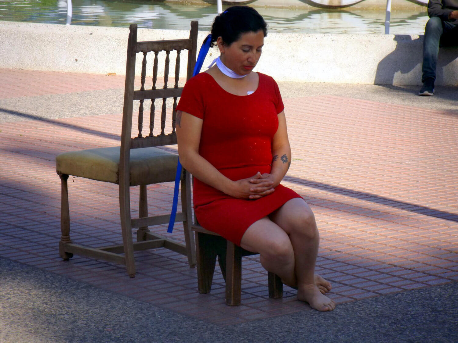 Mujer vestida de rojo, con cintas blancas y azules en su cuello y cabeza, sentada en una silla pequeña, de espaldas a una silla grande desocupada