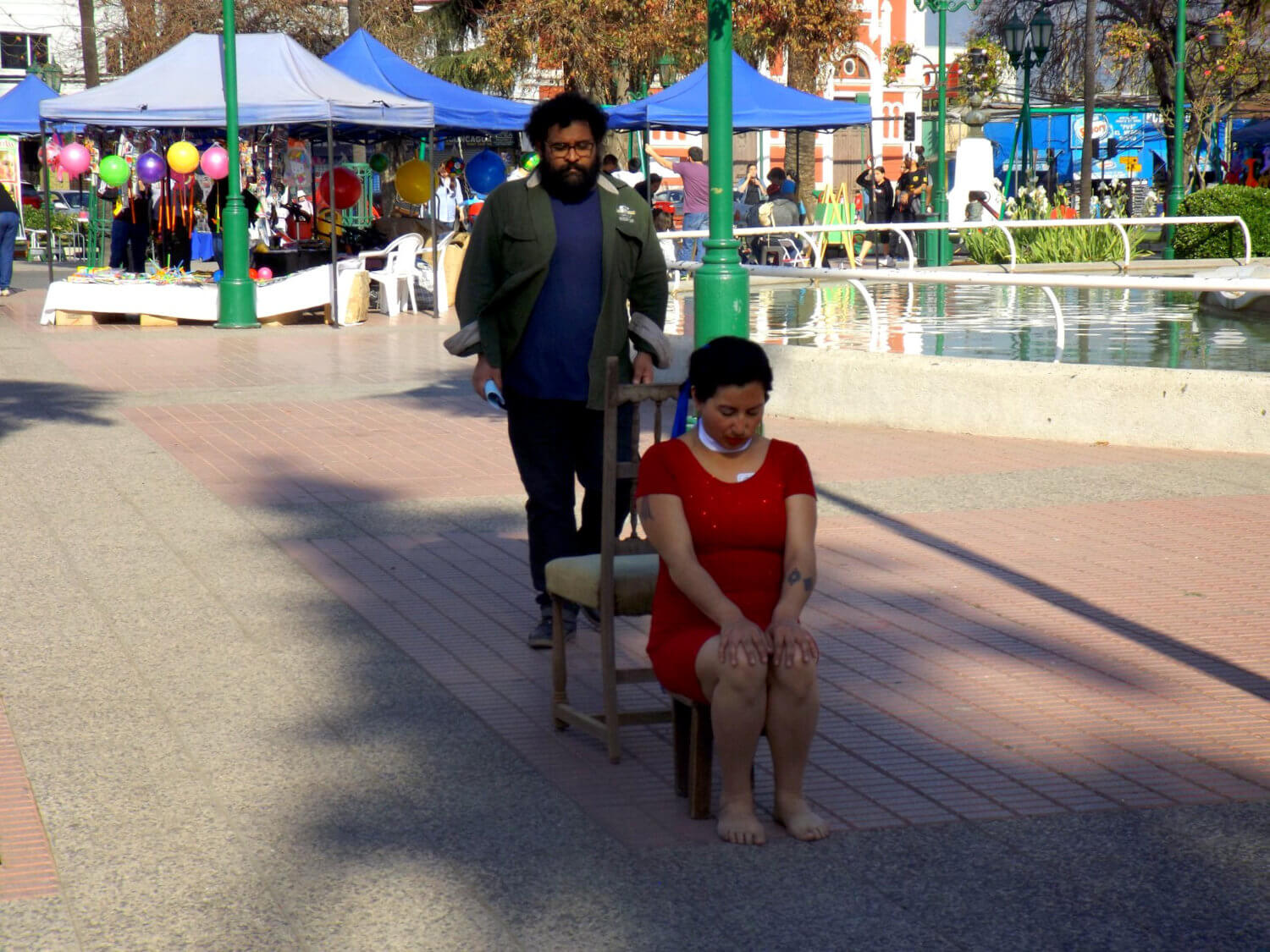 Hombre vestido con polera azul y camisa verde y mujer vestida de rojo, sentada en una silla pequeña, de espaldas a una silla grande desocupada