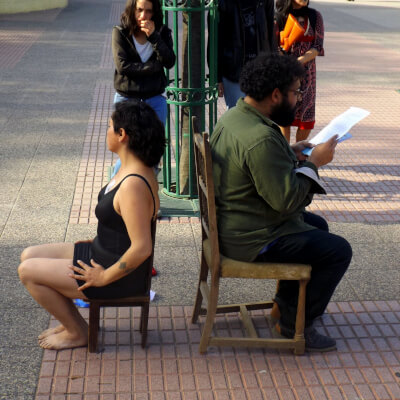 Dos personas, una mujer con blusa y pantalón corto negro, y un hombre que lee un papel, con camisa verde y pantalón azul, sentados cada uno en silla, de espalda el uno al otro, con personas mirando al rededor