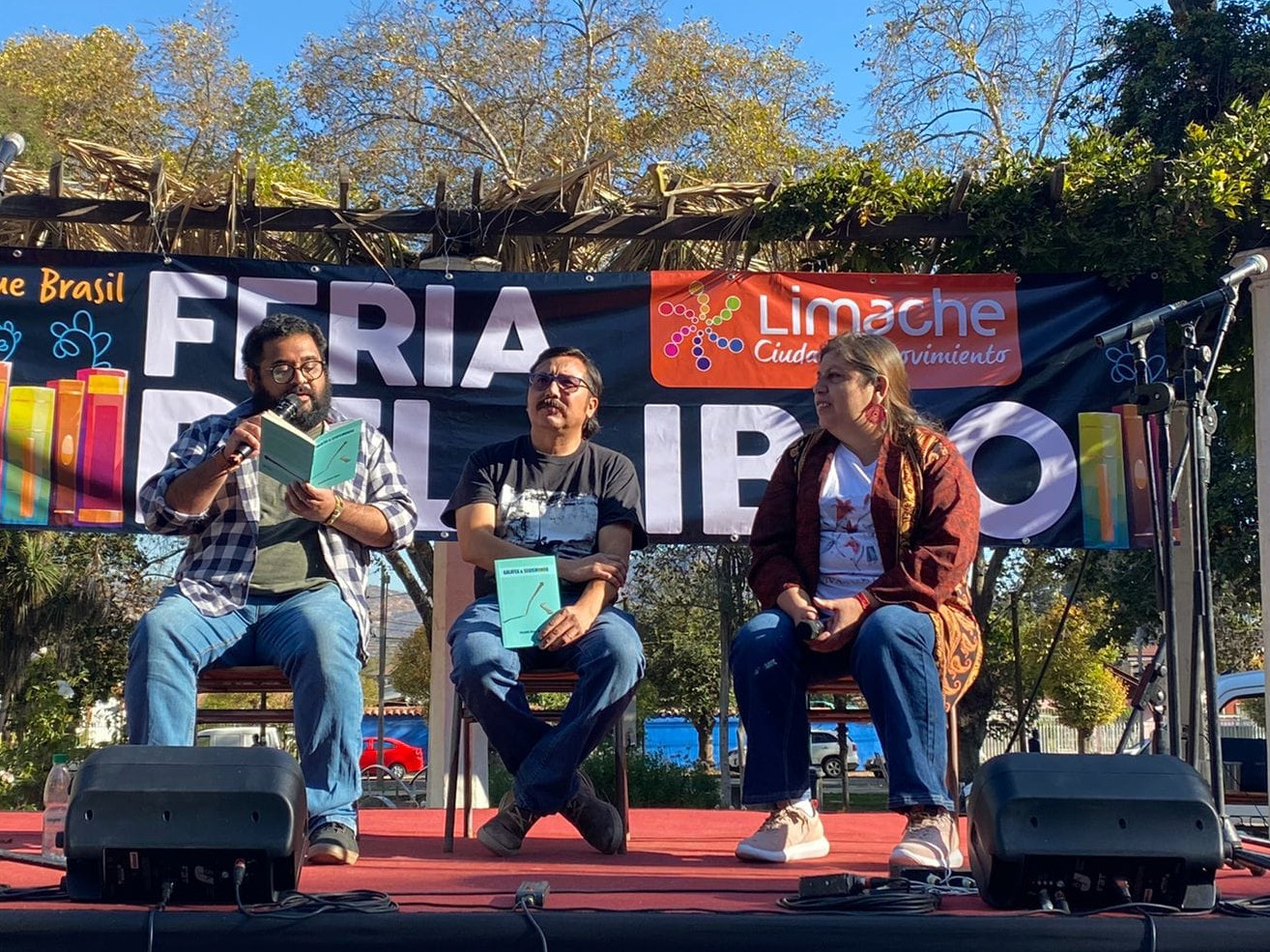 Tres personas sentadas, dos hombres y una mujer, uno leyendo un libro en un escenario de plaza