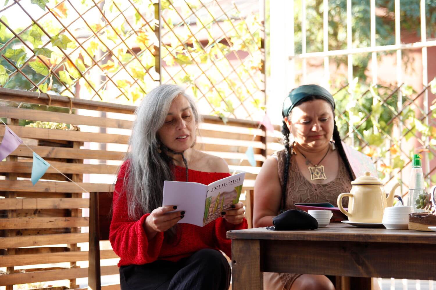 Dos mujeres, una de pelo blanco y vestida con chaleco rojo, leyendo un libro, y otra con cintillo verde y trenzas negras, frente a una mesa con una tetera amarilla